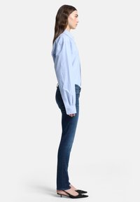 Ljusblå skjorta med långa ärmar, kombinerad med mörkblå jeans och svarta spetsiga klackskor, sedd från sidan.