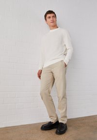 Cremefarbener Strickpullover, beigefarbene Chinos und schwarze Schuhe. Der Pullover hat einen gerippten Saum und Bündchen, während die Chinos eine glatte Textur aufweisen.