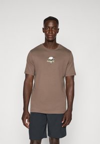 T-shirt en coton marron à manches courtes, avec un col rond et un logo graphique central en jaune et blanc. Associé à un short sombre.