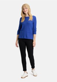 Blauer Pullover mit langen Ärmeln und zwei Vordertaschen, kombiniert mit schwarzen Slim-Fit-Jeans und weißen Sneakern. Einfaches Design, weicher Stoff.