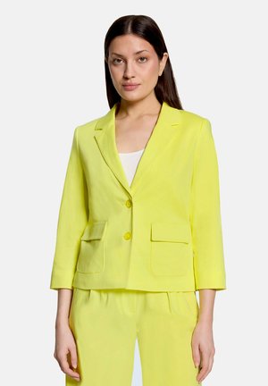 Kvinde iført en klar gul toknaps blazer med forlommer med klap og matchende bukser, stående mod en lys baggrund.