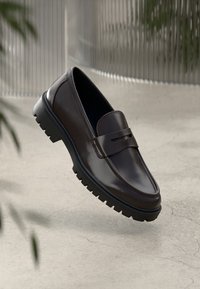 Braune Lederloafers mit glatter Oberfläche, ausgestattet mit einem dekorativen Riemen und einer robusten schwarzen Sohle, die über einer Betonoberfläche schweben.