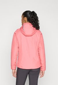 Vrouw met een krullend paardenstaart die een koraalroze hoodie en donkere broeken draagt, van achteren gezien tegen een eenvoudige lichte achtergrond.