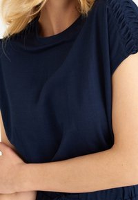 Top de manga corta azul marino hecho de un tejido suave; con detalle fruncido en los hombros y un corte relajado. Vista cercana del escote y las mangas.