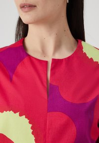 Kortærmet kjole med rød bund, prydet med store lilla og lysegrønne abstrakte former, lavet af blødt stof med en subtil glans.