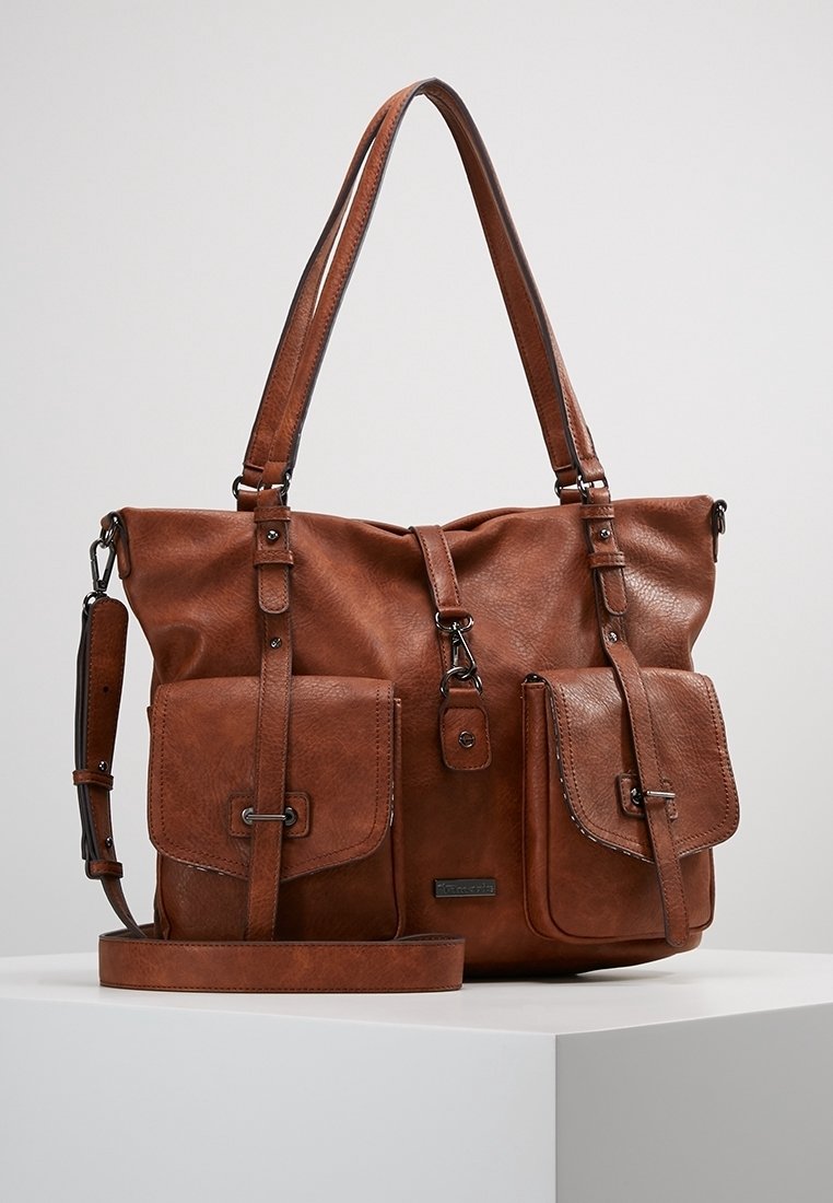 Sac tote en cuir marron avec deux poches avant à boucles, bretelles doubles et une bandoulière ajustable sur une surface blanche.
