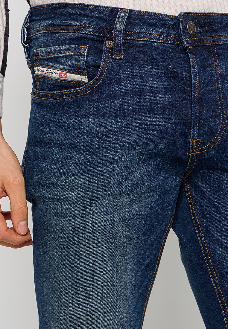 Dunkelblaue Denim-Jeans mit einer Vordertasche, die ein Etikett trägt. Slim Fit, sichtbare Nähte und dezente Farbveränderungen an den Beinen.
