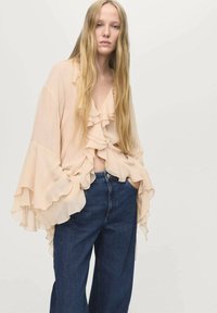 Blouse en chiffon pêche avec un devant froncé, de grandes manches flottantes et un décolleté en V, associé à un jean large bleu marine. Design simple et fluide.