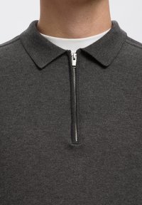 Primo piano di una persona che indossa una camicia grigio scuro con colletto e una cerniera verticale argentata parzialmente slacciata sopra una maglietta bianca.
