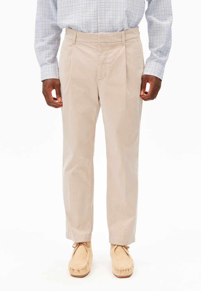 Beige plissierte Hose aus leichtem Stoff, mit Reißverschluss, Seitentaschen und schmal zulaufender Passform, kombiniert mit tanfarbenen Mokassins.