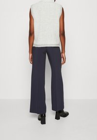 Persona in piedi di spalle con indosso un top bianco senza maniche, pantaloni larghi blu navy e stivaletti neri con tacco su uno sfondo semplice.