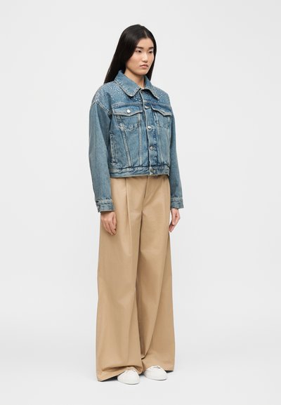 Νέα γυναίκα φορώντας ένα μπλε τζιν μπουφάν με κεντήματα, μπεζ παντελόνι φαρδύ στο κάτω μέρος και λευκά αθλητικά παπούτσια, στέκεται μπροστά σε ένα απλό φωτεινό φόντο.