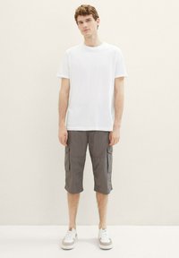Weißes Baumwoll-T-Shirt kombiniert mit grauen Cargo-Shorts mit Seitenfächern. Das Model trägt weiße Sneakers mit beigen Akzenten. Neutraler Hintergrund.