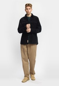 Zwarte corduroy overhemdjas met twee borstzakken, knoopsluiting en een gestructureerde afwerking, gecombineerd met lichtbeige broeken en tan sneakers.