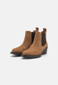 Botines de ante marrón con punta redonda, paneles laterales elásticos y un ligero tacón; textura suave con tiradores para facilitar el calzado.
