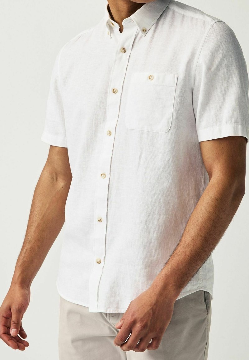 Homme portant une chemise blanche à manches courtes avec une poche poitrine et un pantalon de couleur claire sur un fond uni.