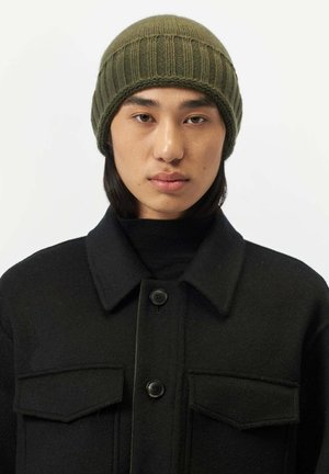 Young person with straight black hair wears olive green knit beanie and black buttoned coat with chest pockets, facing forward.