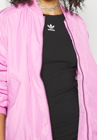 Rosa lätt bomberjacka med dragkedja fram, ribbade ärmar och en slät yta. Svart linne med vitt Adidas-logga under.