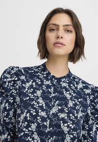 Navy blue blouse with white floral pattern, puffed sleeves, and button front. Features a collar and textured fabric.
