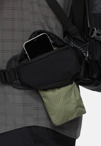 Bolsa de cintura de tela negra con cremallera, que contiene un teléfono inteligente, sujeta a una camisa de cuadros gris; bolsa verde texturizada visible debajo.
