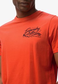 Camiseta de algodón roja con un gráfico negro de montañas y árboles, con el texto "Superdry" y "Confía en lo Mejor" impreso en el pecho.