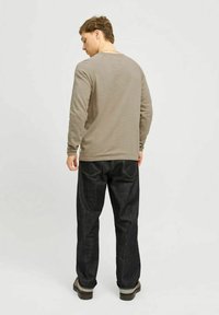 Camicia a maniche lunghe marrone chiaro e jeans di jeans scuri, con una vestibilità comoda e dettagli di cucitura chiara, in piedi su uno sfondo neutro.