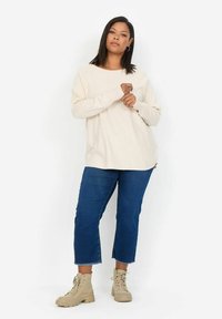 Beige langärmliges Oberteil mit lockerer Passform, kombiniert mit verkürzten blauen Jeans und braunen Schnürstiefeln. Schlichtes Design, glatte Textur.