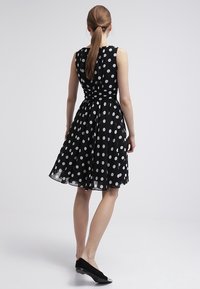 Robe sans manches noire avec des pois blancs, taille froncée, jupe évasée et tissu transparent. Complétée par des chaussures plates noires.