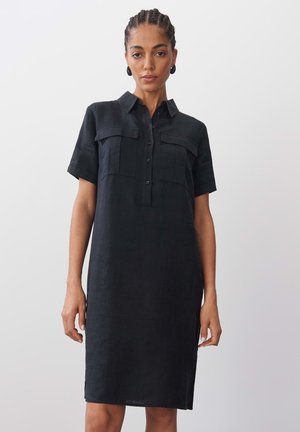 Vestito di lino nero con collo a camicia, maniche corte, doppie tasche frontali e vestibilità ampia. Presenta una chiusura con bottoni e fessure laterali.