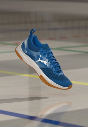 Blauer Sportschuh mit weißem Puma-Logo, Mesh-Obermaterial, weißer Mittelsohle und Gummiaußensohle, der über dem Boden eines Hallensportplatzes schwebt.