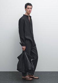 Zwarte langwerpige blouse met knopen, gecombineerd met donkere jeans en zwarte slippers. Een grote zwarte leren tote bag wordt in één hand vastgehouden.