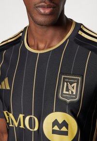 Maglia da calcio nera con strisce dorate, scollatura rotonda e dettagli dorati. Presenta il logo del Los Angeles Football Club e il branding dello sponsor.