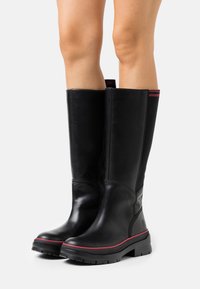 Bottes noires en cuir montant jusqu'au genou avec une semelle en caoutchouc, des accents rouges et des panneaux élastiques pour plus de flexibilité. Texture lisse avec un design à bout rond.