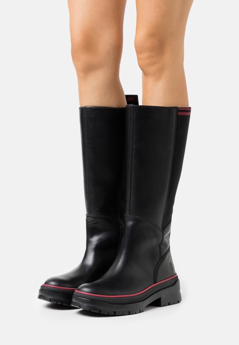 Bottes noires en cuir montant jusqu'au genou avec une semelle en caoutchouc, des accents rouges et des panneaux élastiques pour plus de flexibilité. Texture lisse avec un design à bout rond.