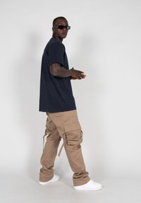 Marinemariner kortærmet t-shirt, beige cargo bukser med sidelommer, hvide sneakers og mørke solbriller, poseret mod en neutral baggrund.