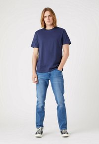 T-shirt in cotone blu navy con colletto a girocollo, abbinato a jeans azzurri e scarpe a righe bianche e nere, in piedi contro uno sfondo chiaro.