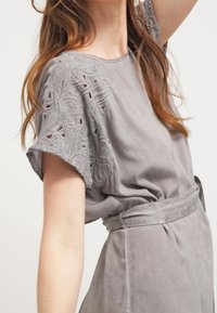 Harmaa mokkatakkimekko, jossa on lyhyet hihat ja kirjailtuja kukkaleikkauksia, vyötäröllä vyö ja rento istuvuus. Tasainen pinta, jossa on pehmeä kiilto.