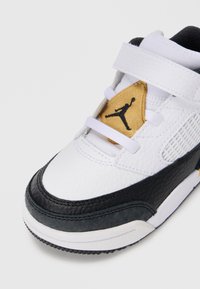 Adidași albi cu accente negre, material din piele texturată, design unghiular și un etichetă aurie cu un logo Jumpman negru.