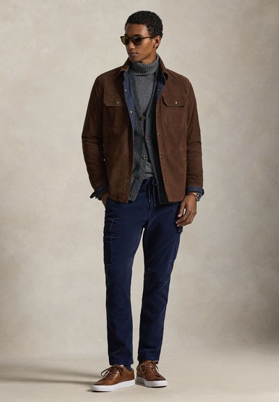 Καφέ σουέντ μπουφάν πάνω από γκρι πουλόβερ με ζιβάγκο και μπλε πουκάμισο, συνδυασμένο με μπλε σκούρο παντελόνι cargo και καφέ αθλητικά παπούτσια, τονισμένο με γυαλιά ηλίου.