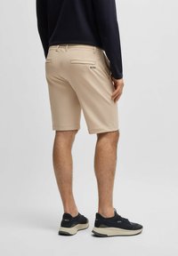 Pantaloni corti beige da uomo, vestibilità su misura, realizzati in tessuto leggero, con tasche posteriori e un piccolo logo. Indossati con scarpe da ginnastica nere.