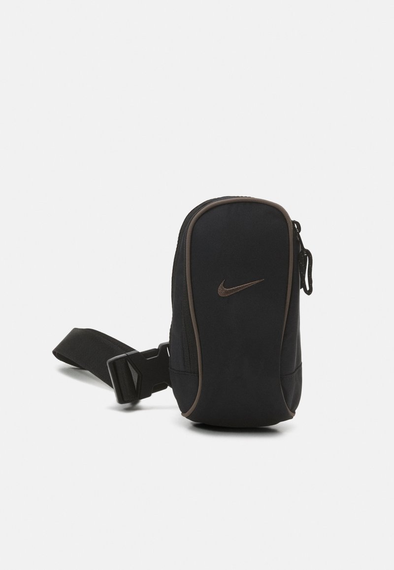 Černá textilní crossbody taška s hladkým povrchem, na zip, a tmavě hnědým akcentem podél okrajů. Obsahuje malé logo Nike.