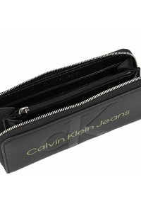 Portafoglio in pelle nera con chiusura a zip, dotato di logo "CK" in rilievo e "Calvin Klein Jeans" in verde. L'interno presenta numerosi scomparti.