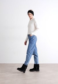 Model draagt een witte gebreide trui, lichtblauwe jeans en zwarte enkellaarsjes. Accessoires bestaan uit een zwarte haarband. Eenvoudige achtergrond.