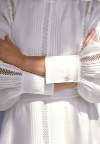 Robe en tissu blanc texturé avec des détails plissés, dotée de manches longues, de fermetures à boutons et d'accents en dentelle complexes le long des bras.