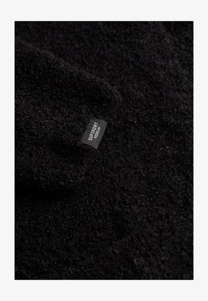 Zbliżenie na czarną teksturowaną tkaninę z małą czarną metką z napisem "SUPERDRY PREMIUM" przyszytą do niej.