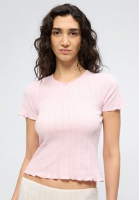 Γυναίκα με σκούρα, σγουρά μαλλιά, που φοράει ανοιχτό ροζ κοντομάνικο ριμπ τοπ και ανοιχτόχρωμο παντελόνι, στέκεται μπροστά σε απλό φόντο.