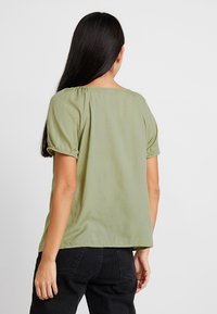 Blusa de manga corta, verde oliva, con un corte holgado, que presenta detalles fruncidos en los hombros y un escote redondeado.