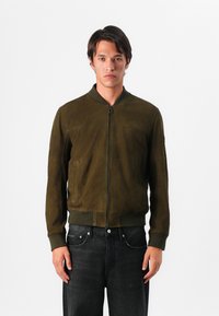 Λάδι πράσινη bomber τζάκετ από σουέτ, με μπροστινό φερμουάρ, ριμπ μανσέτες και δύο πλαϊνές τσέπες, σε συνδυασμό με σκούρα τζιν.