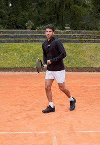Um jogador de ténis masculino, vestindo um capuz preto e calções brancos, está de pé numa quadra de terra batida, segurando uma raquete de ténis, com sapatos pretos e meias de cano curto.