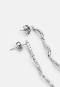 Brincos de metal prateado com um design de corrente interligada, apresentando formas suaves e onduladas, e um fecho traseiro de pino seguro.